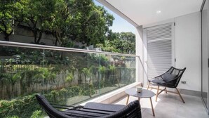 A balcony with a glass railing, two black wicker chairs, a small round table, and a view of a garden with trees and a water feature. - H401 | top-Rated Cali Retreat 2BR w/ Balcony & Pool 24/7 Security + Free Parking (Cali)