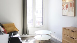 A room with a sofa, a round table, a wooden dresser, and a window with curtains. - Charming T1 at the gates of Paris (Asnières-sur-Seine)