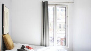 A bedroom with a bed, a window with curtains, a view of buildings outside, and a wall-mounted picture. - Charming T1 at the gates of Paris (Asnières-sur-Seine)