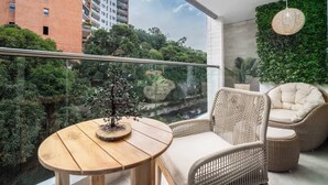 A balcony with a wooden table, wicker chairs, a potted bonsai tree, and a view of a cityscape and greenery. - H702 - Amazing Balcony | Pool | Gym | Near Granada (Cali)