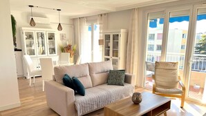 A modern living room with a sofa, a wooden coffee table, and a chair. There is a dining area with a white table and chairs. The room has large windows with curtains, and a balcony is visible through the windows.
