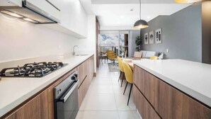 A modern kitchen with a gas stove, oven, and dishwasher, a dining area with yellow chairs, and a view of the city through large windows. - H706 - Stylish 2BR / Perfect Views & Rooftop Pool (Cali)