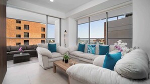 Una sala de estar moderna con un amplio sofá, una mesa de centro y vistas a un edificio de ladrillo a través de grandes ventanales.