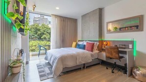 Ein modernes Schlafzimmer mit Bett, Schreibtisch und Stuhl, ein Balkon mit Vorhängen und Blick ins Grüne.