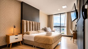 A modern hotel room with a large bed, a desk, a chair, a TV, and a view of the city. - RM 1102 | Luxurious Suite | Pool | Gym | Security (Cali)