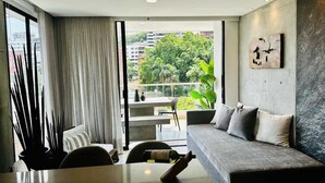 Un salon moderne avec un canapé, une table à manger et une vue sur la verdure et les bâtiments, grâce à de grandes fenêtres.