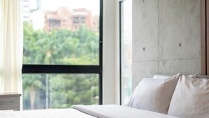 Een slaapkamer met een groot raam, een bed met witte lakens en een betonnen muur.
