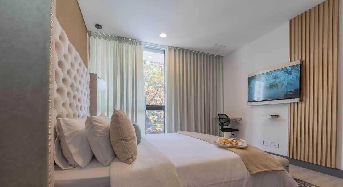 A modern hotel room with a large bed, a flat-screen TV mounted on the wall, and a view of the outdoors through the window.