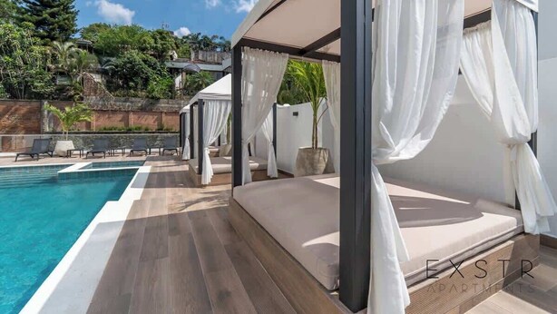 Un dormitorio moderno al aire libre con piscina, cortinas blancas y vistas de la vegetación circundante.