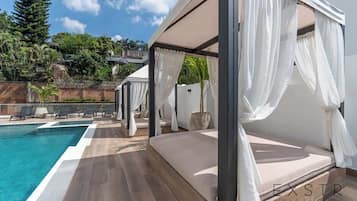 A modern outdoor bedroom with a pool, white curtains, and a view of the surrounding greenery.
