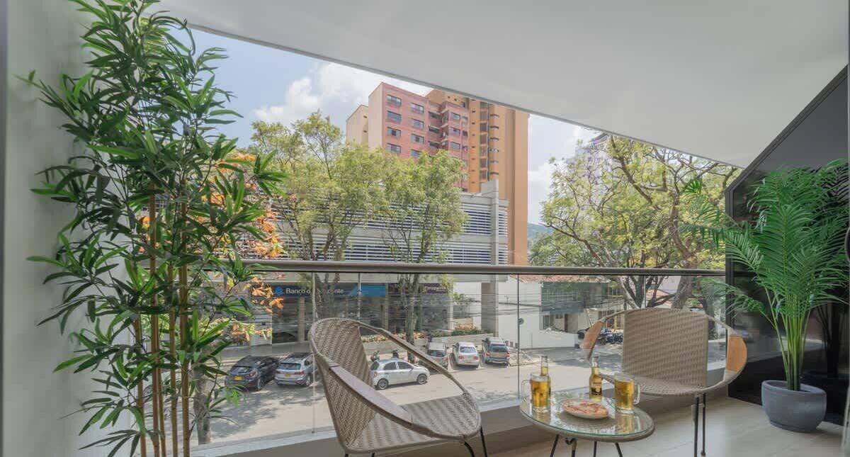 A balcony with a view of a cityscape, including a tall building and parked cars. There are two wicker chairs and a table with a bottle and glass.