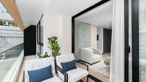 A modern living room with a balcony, a large plant, and a view of the city. - RM 208 | Gorgeous 1-Bed | Parking | Pool | Sauna (Cali)