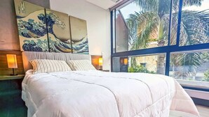 A hotel room with a large bed, a painting of waves on the wall, and a view of palm trees through the window. - Dazzling Brand-New 1 BD w Terrace & Pool (Cali)