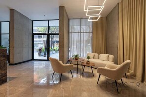 Ein modernes Interieur mit einer Sofagarnitur, zwei Sesseln und einem kleinen Tisch. Ein großes Fenster mit Vorhängen, eine Sichtbetonsäule und ein Blick ins Freie.