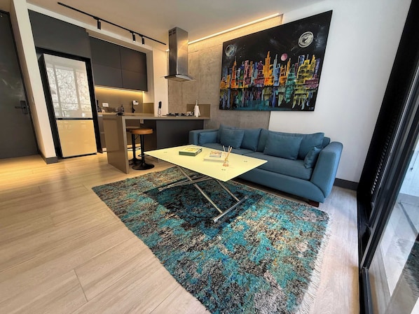 A modern living space with a sofa, a coffee table, a kitchen area, and a painting on the wall. - RM 308 | Luxury | Pool & Parking | Santa Teresita (Cali)