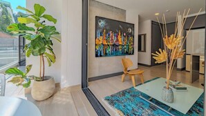 A modern interior with a large window, a colorful cityscape painting, a potted plant, a wooden chair, and a glass table with a blue rug. - RM 308 | Luxury | Pool & Parking | Santa Teresita (Cali)