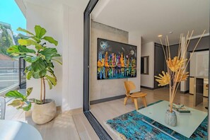 Ein modernes Interieur mit einem großen Fenster, einem farbenfrohen Stadtbildgemälde, einer Topfpflanze, einem Holzstuhl und einem Glastisch mit blauem Teppich.
