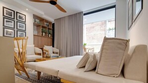 A modern hotel room with a bed, two chairs, a small table, a bookshelf, and a large window.