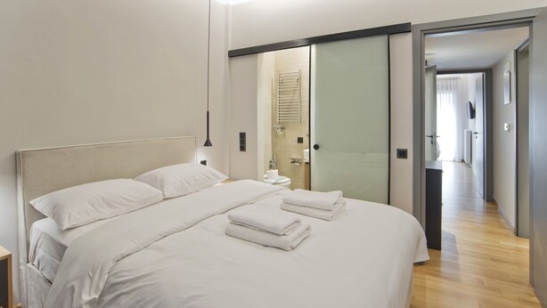 A modern hotel room with a large bed, a bedside table, and a bathroom visible through a glass door. - Stylish Ilisia Stay Luxury Fully Reno 2BR, 2BA Apt (Athens)
