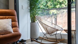 A modern living room with a brown leather sofa, a woven rocking chair, a potted plant, and a view of greenery outside.