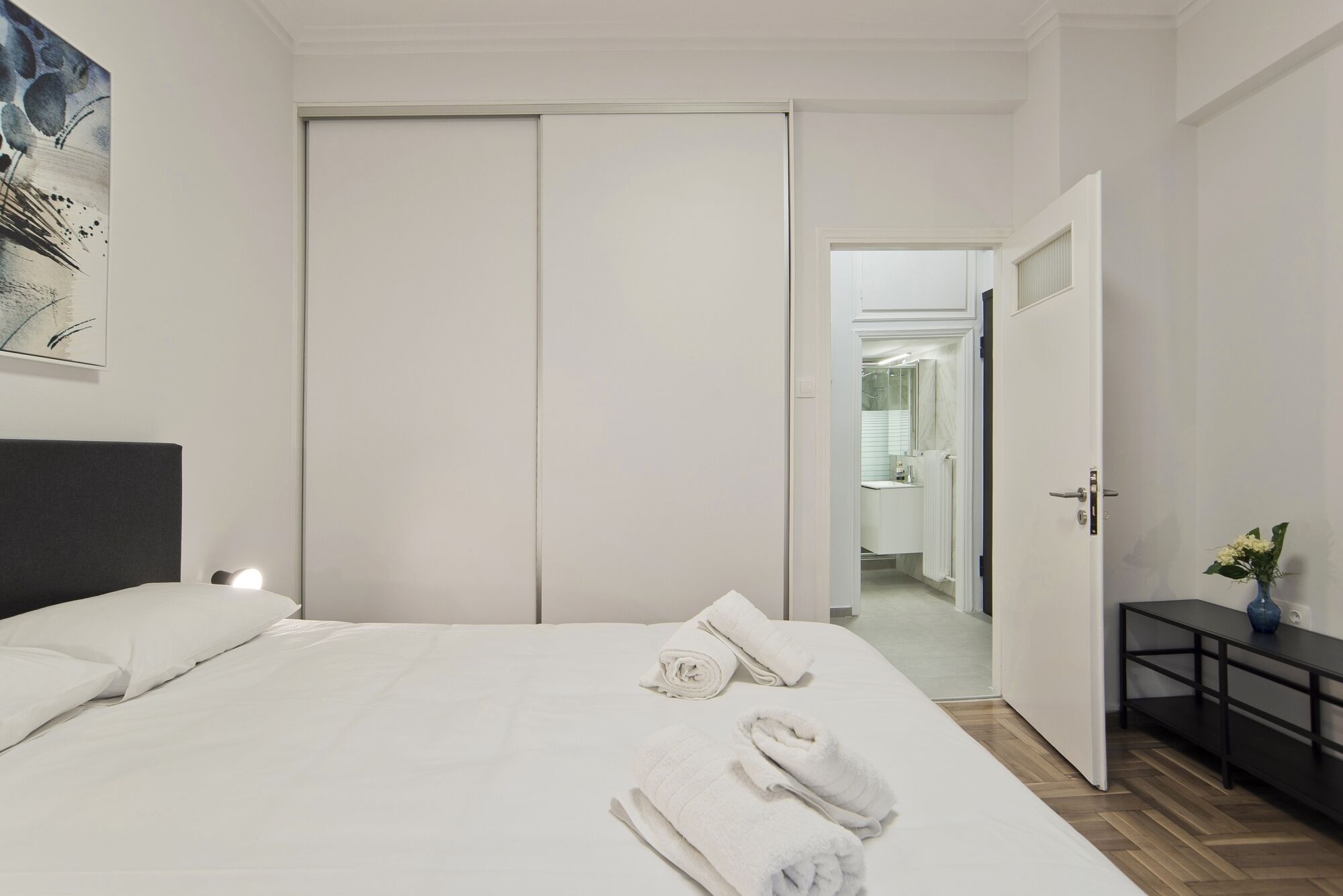 Une chambre d’hôtel comprenant un lit, une table de chevet, une commode, une porte donnant sur une salle de bain et un tableau accroché au mur.