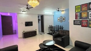 Un salon moderne avec un canapé en cuir noir, une table basse et un lustre. On y trouve également un coin repas avec une table et des chaises, ainsi qu’un téléviseur fixé au mur.