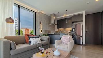 Una sala de estar moderna con sofá, sillón y mesa de centro. Cuenta con una zona de cocina equipada con refrigerador y armarios.