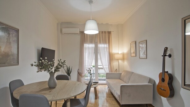 A modern living room with a round wooden table, grey chairs, a white sofa, a guitar, and a large window with sheer curtains.
