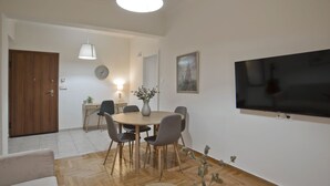A modern living room with a round dining table, grey chairs, a flat-screen TV mounted on the wall, and a wooden door.