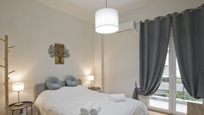 A bedroom with a bed, two bedside lamps, a wall art of a tree, a window with curtains, and a balcony view.
