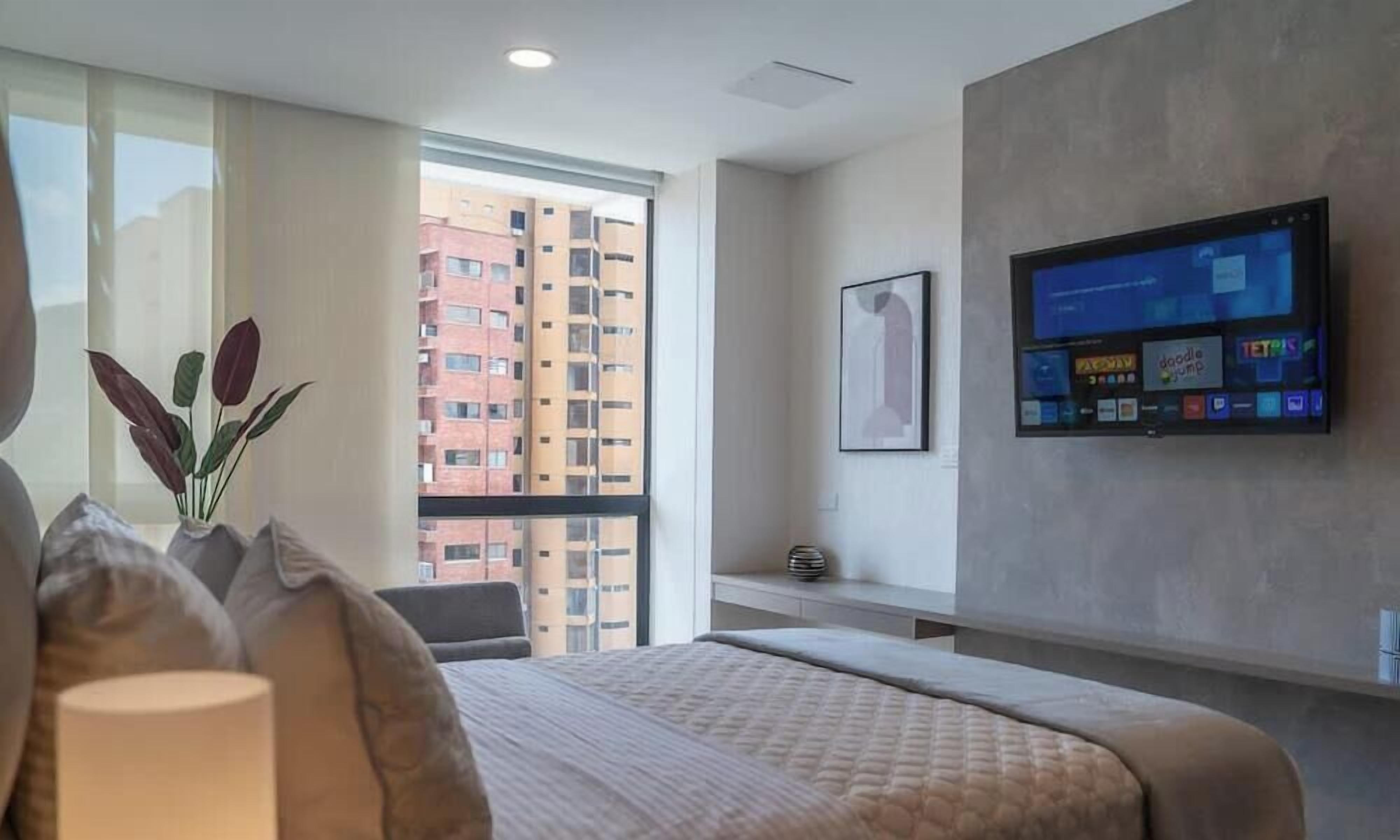 Habitación de hotel con una cama grande, un televisor montado en la pared, una ventana con vistas a rascacielos y un florero con plantas en el alféizar.