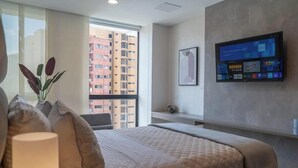 A hotel room with a large bed, a television mounted on the wall, a window with a view of high-rise buildings, and a vase with plants on the windowsill. - RM902| LuxeHigh- Hideaway – Pool, Sauna & Security (Cali)