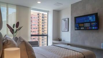 Habitación de hotel con una cama grande, un televisor montado en la pared, una ventana con vistas a rascacielos y un florero con plantas en el alféizar.