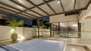 An indoor recreational area with a ping pong table, glass walls, and a ceiling with exposed beams. - RM902| LuxeHigh- Hideaway – Pool, Sauna & Security (Cali)