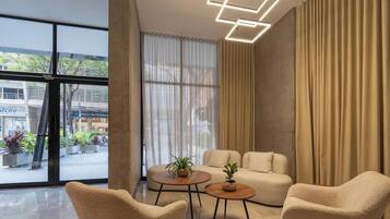 Una sala moderna con un sofá, dos sillones y una mesa de centro. Hay ventanas amplias con cortinas, vistas a la calle y una lámpara de techo con diseño geométrico.