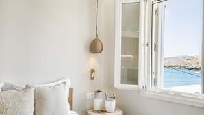 A bedroom with a bed, a bedside table with decorative items, a wall-mounted light, and a window with a view of the sea. - Kythnos Blue - Suite 4 Luxurious, Sea Front (Kythnos)
