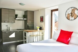 A modern hotel room with a kitchen area, a dining table, and a bed with a red pillow.