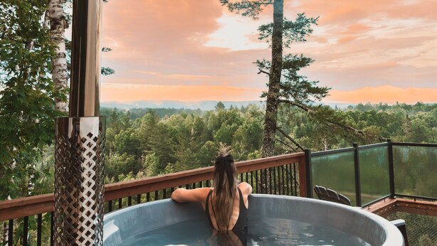Una persona en un jacuzzi en una terraza con vista a un bosque y montañas.