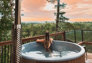 Eine Person in einer Badewanne auf einer Terrasse mit Blick auf einen Wald und Berge.