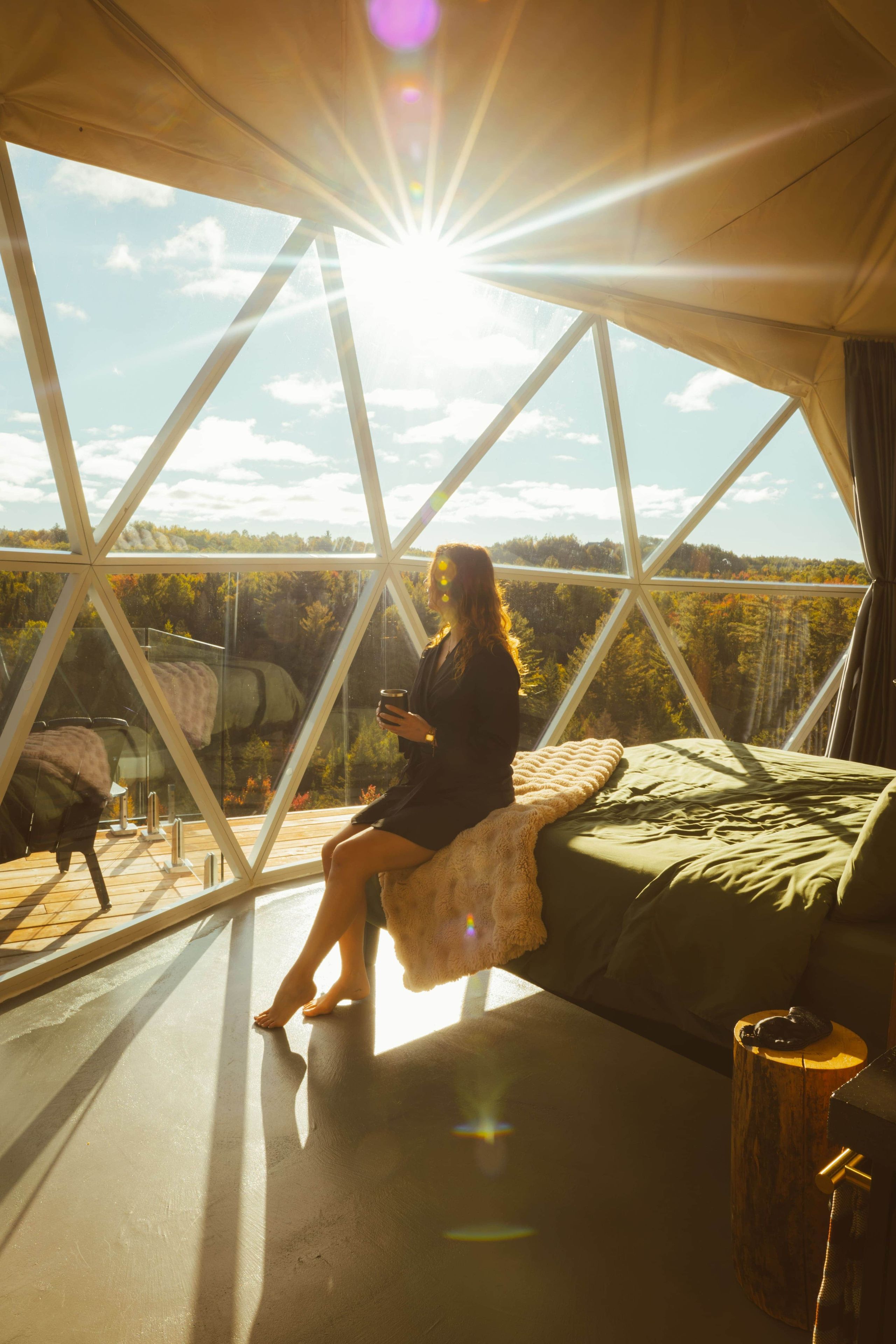 Una persona sentada en una cama en una habitación con ventanas amplias, vista a árboles y suelo de madera.