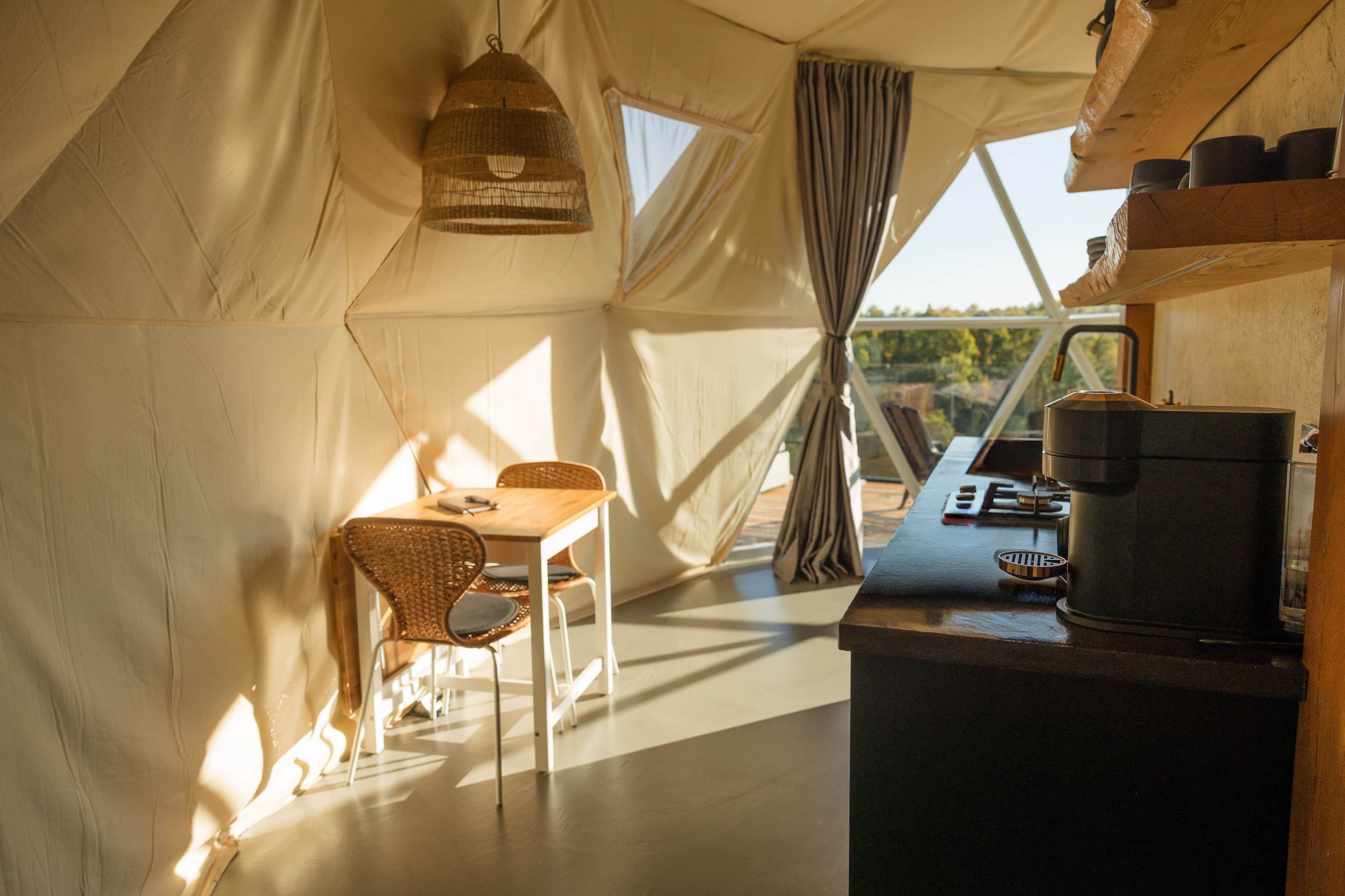 Acomodação em tenda com área de refeições, uma mesa, cadeiras, luminária pendente e cozinha equipada com cafeteira.