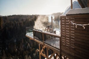 A wooden deck with a hot tub surrounded by snow-covered trees.