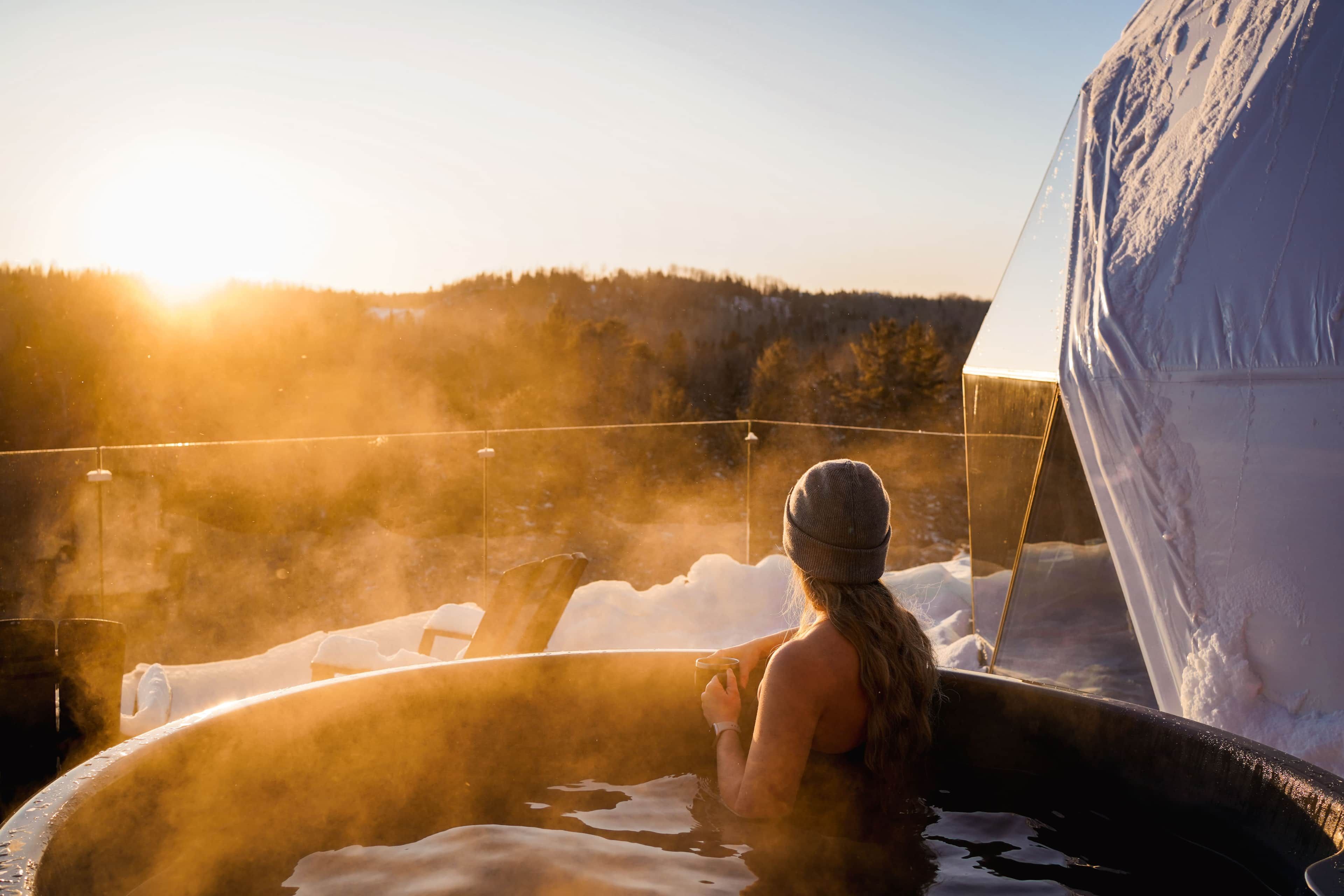 Une personne dans un bain à remous, avec vue sur un paysage enneigé et une tente en arrière-plan.