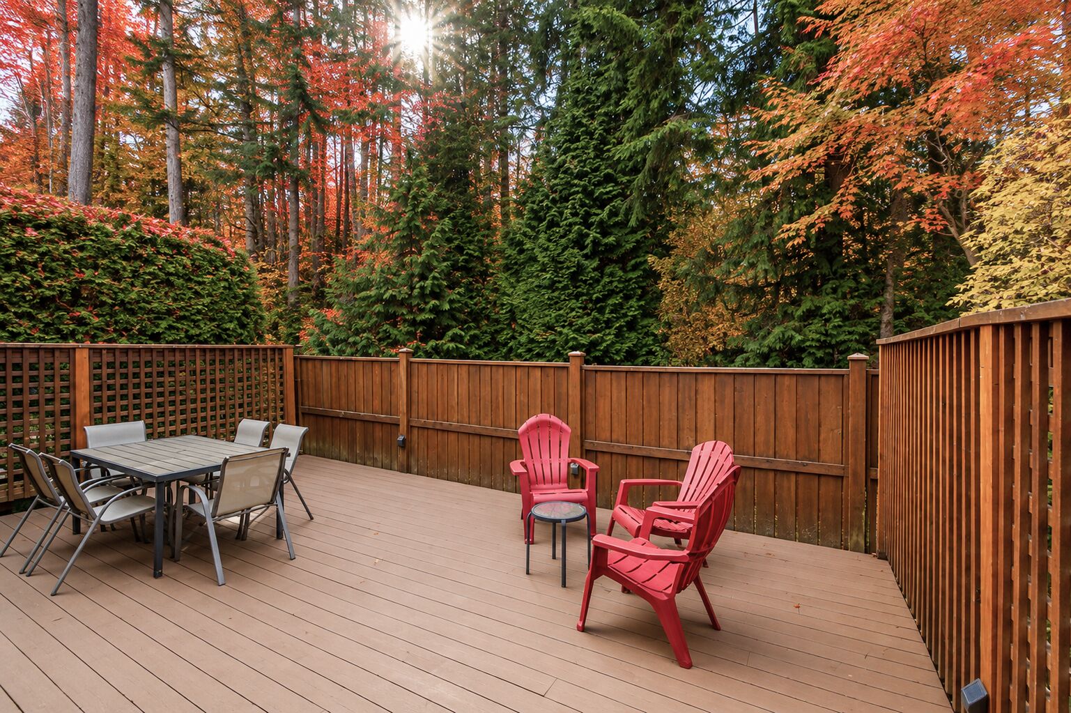 An outdoor deck with a wooden fence, a table with chairs, and red Adirondack chairs.