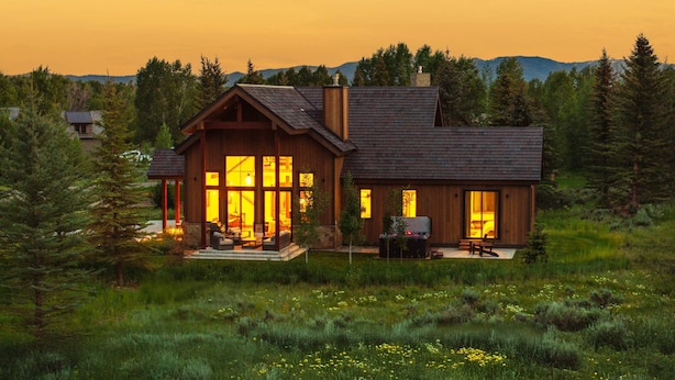 A wooden cabin with large windows, a deck, and a grill area, surrounded by a forested landscape.