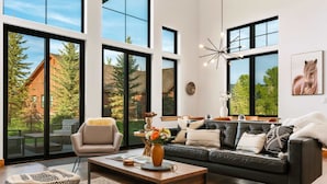 A modern living room with a large window, a black leather sofa, a wooden coffee table, and a patterned ottoman.