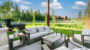 An outdoor seating area with a sofa, coffee table, and chairs, overlooking a grassy field and a wooden cabin.