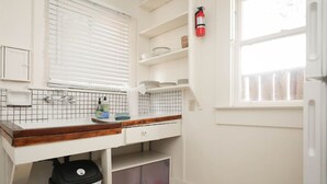 Una cocina pequeña con encimera blanca, ventana con persianas, extintor y cubo de basura.