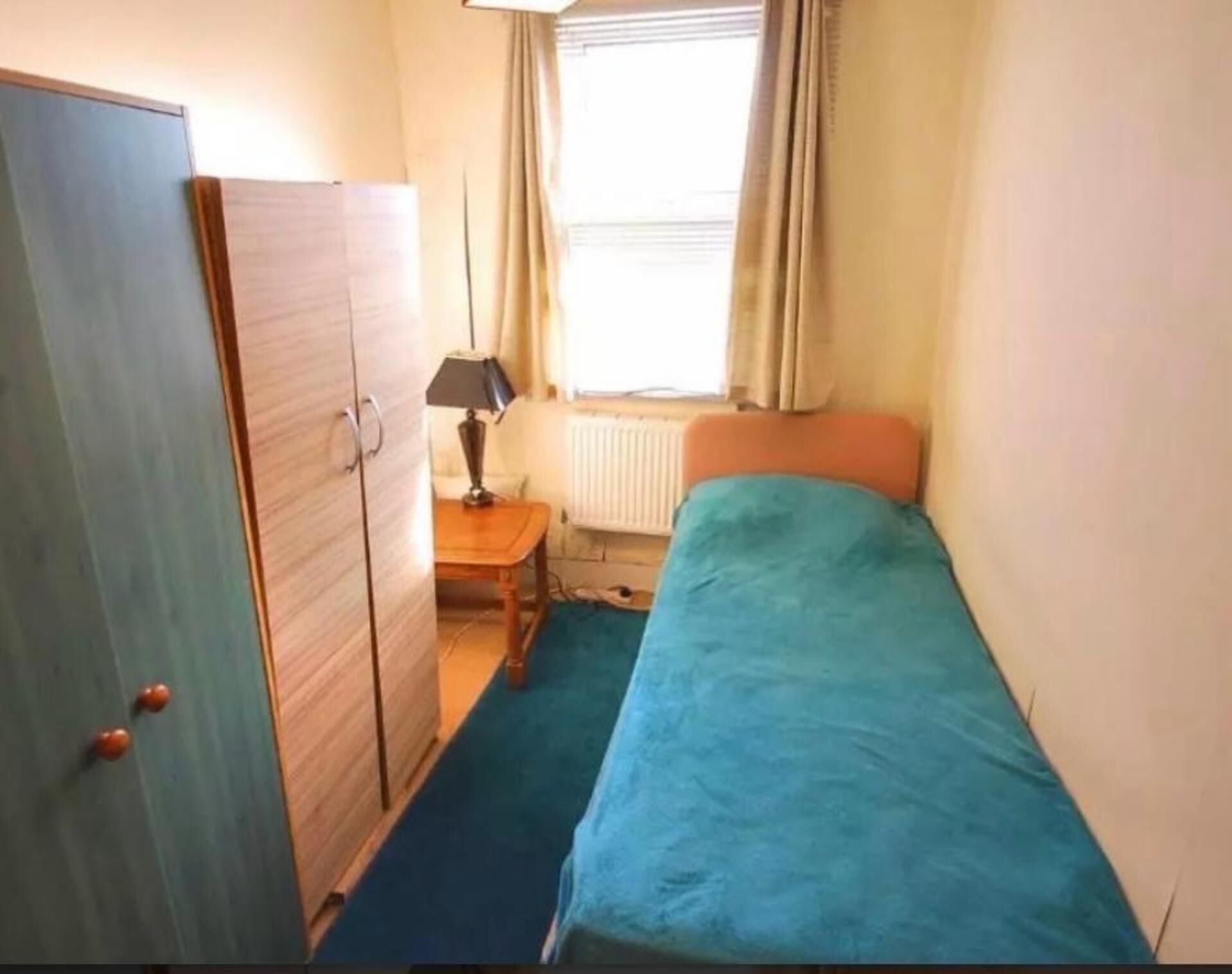 A small, single-bed room with a wooden wardrobe, a small table with a lamp, and a window with curtains.