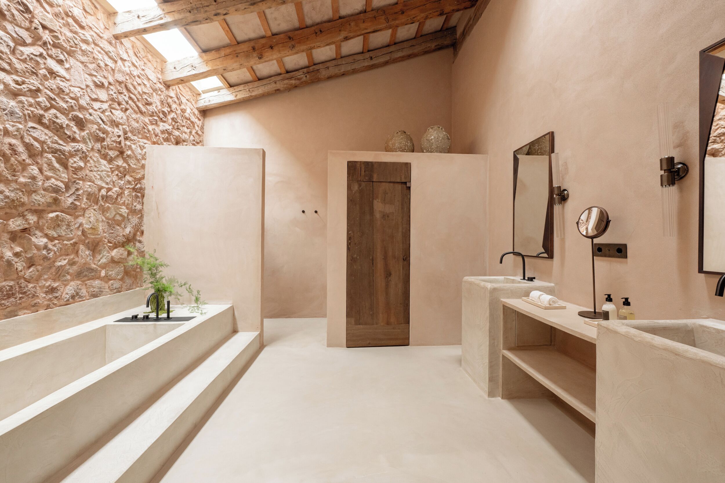 Un baño con paredes de piedra, puerta de madera y dos lavamanos con espejos.